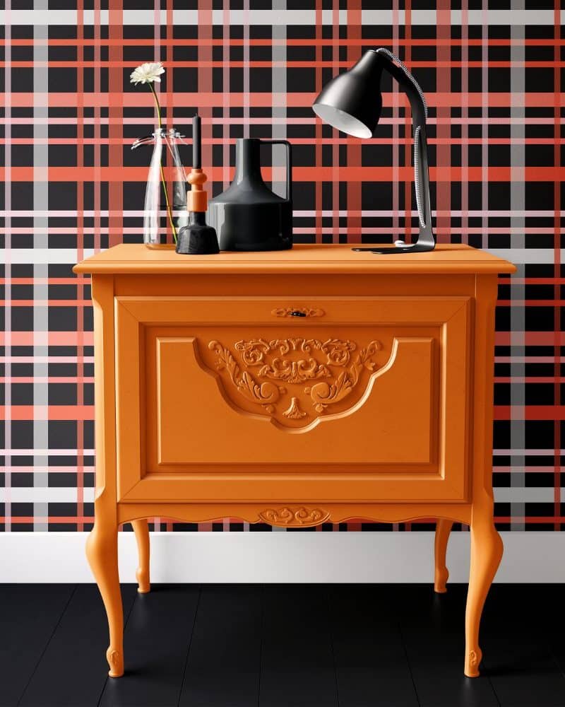 Orange wooden cabinet with ornate detailing, holding a lamp and decorative objects, against a black and coral plaid wall.