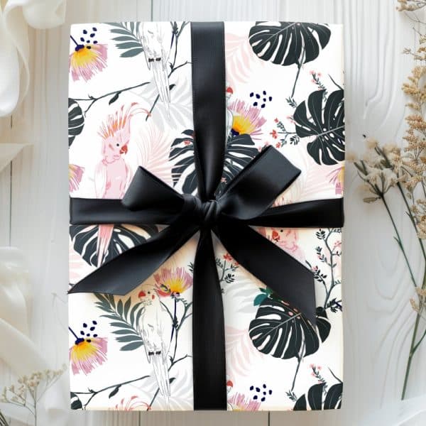 A Gift Wrapped In White Paper Featuring Pink And White Parrots And Tropical Leaves, Tied With A Black Ribbon.