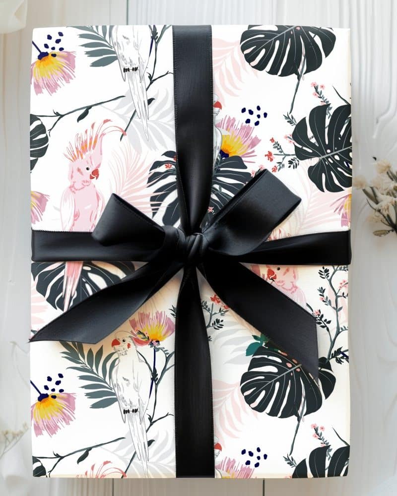 A gift wrapped in white paper featuring pink and white parrots and tropical leaves, tied with a black ribbon.