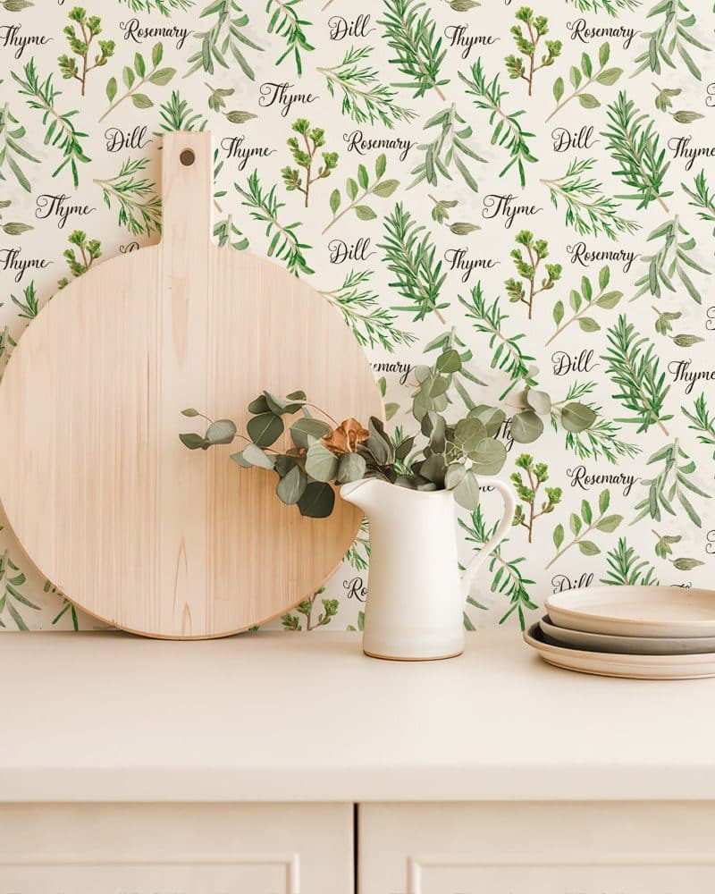 A kitchen counter with a wooden cutting board, a white creamer, and stacked plates against a backdrop of herb-themed wallpaper.