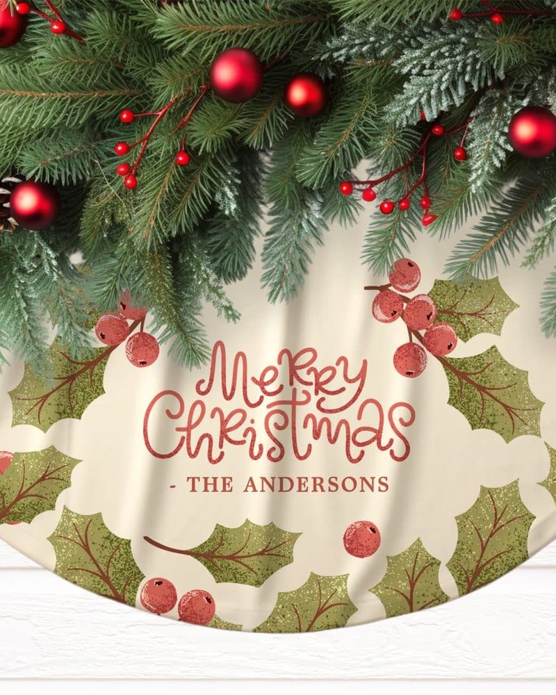 A cream-colored Christmas tree skirt with a Merry Christmas message and The Andersons family name, decorated with holly leaves and berries, is topped with a garland of pine boughs and red ornaments.