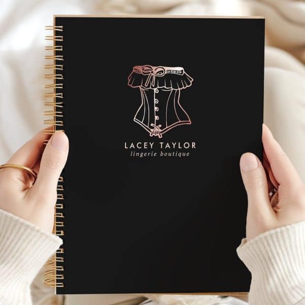 Hands Hold A Black Spiral Notebook Featuring A Rose Gold Logo Of A Corset And The Text Lacey Taylor Lingerie Boutique.