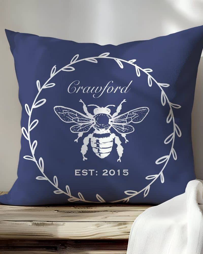 Navy blue throw pillow with a white bee illustration, wreath, and the name Crawford.