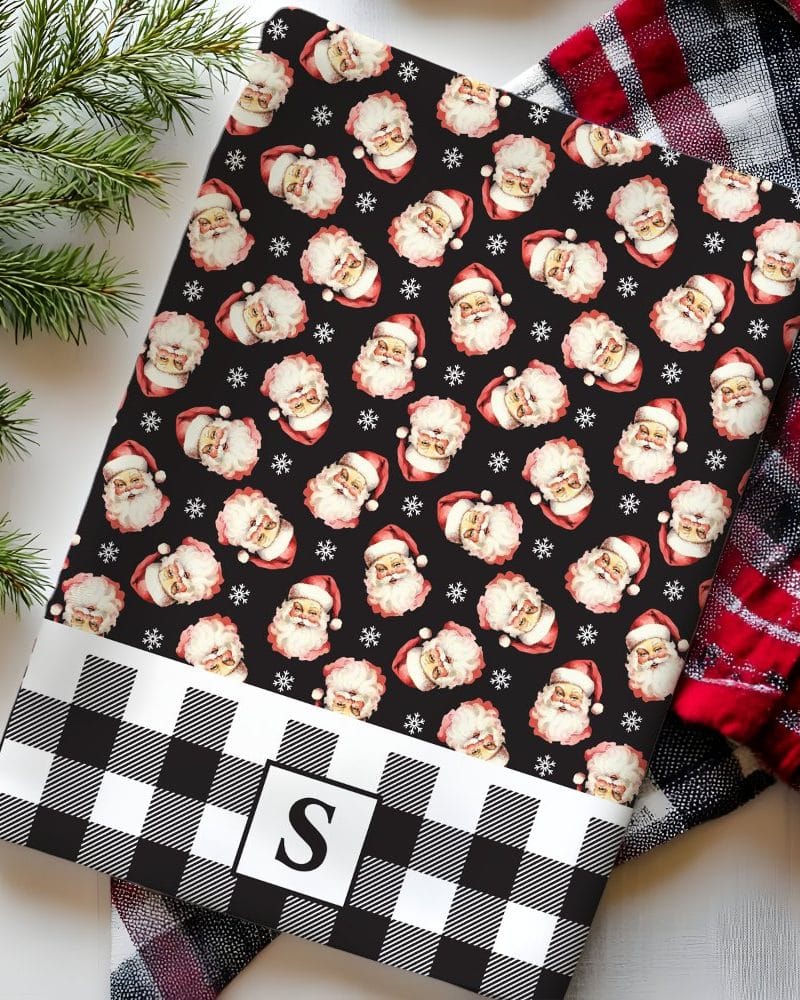 A black and white buffalo plaid Christmas gift wrap featuring a repeating pattern of Santa Claus faces and snowflakes. A monogrammed initial S is in the center of the plaid bottom.