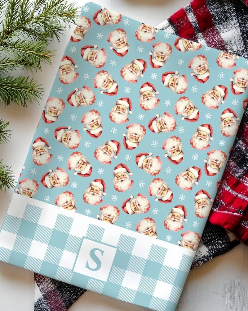 A light blue and white gingham-patterned wrapping paper featuring a repeating pattern of vintage Santa Claus faces. The bottom has a monogrammed S.