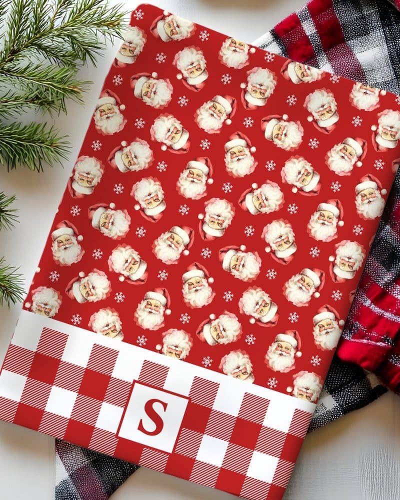 Red and white buffalo plaid placemat with a repeating pattern of Santa Claus faces and snowflakes above the plaid and a monogrammed S in the center of the plaid.