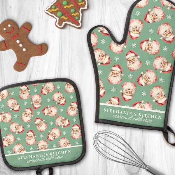 A Pair Of Personalized Christmas Oven Mitts And Pot Holders. The Green Fabric Features A Repeating Pattern Of Santa Claus Faces And Snowflakes. The Mitts Are Personalized With The Text &Quot;Stephanie'S Kitchen Seasoned With Love&Quot;.