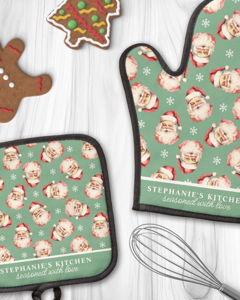 A pair of personalized Christmas oven mitts and pot holders. The green fabric features a repeating pattern of Santa Claus faces and snowflakes. The mitts are personalized with the text "Stephanie's Kitchen seasoned with love".