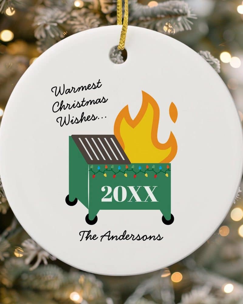 Round Christmas ornament with a dumpster fire illustration, Christmas lights, and the text Warmest Christmas Wishes, 20XX, and The Andersons.