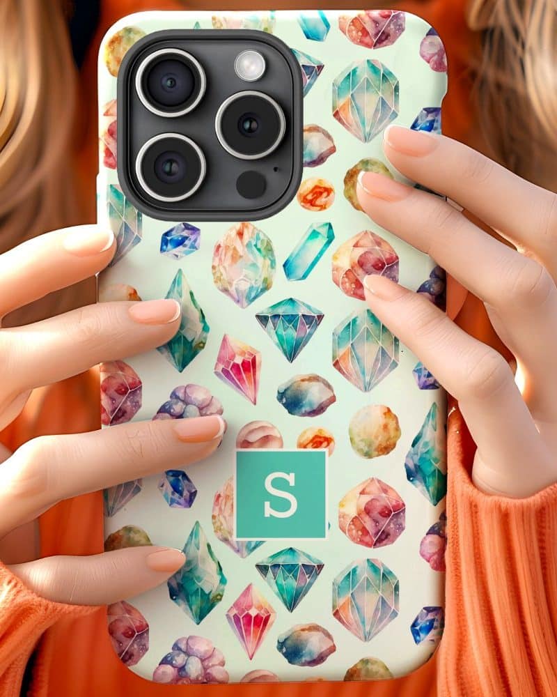 Woman's hands holding a phone case featuring a watercolor pattern of colorful gemstones and a teal initial S.