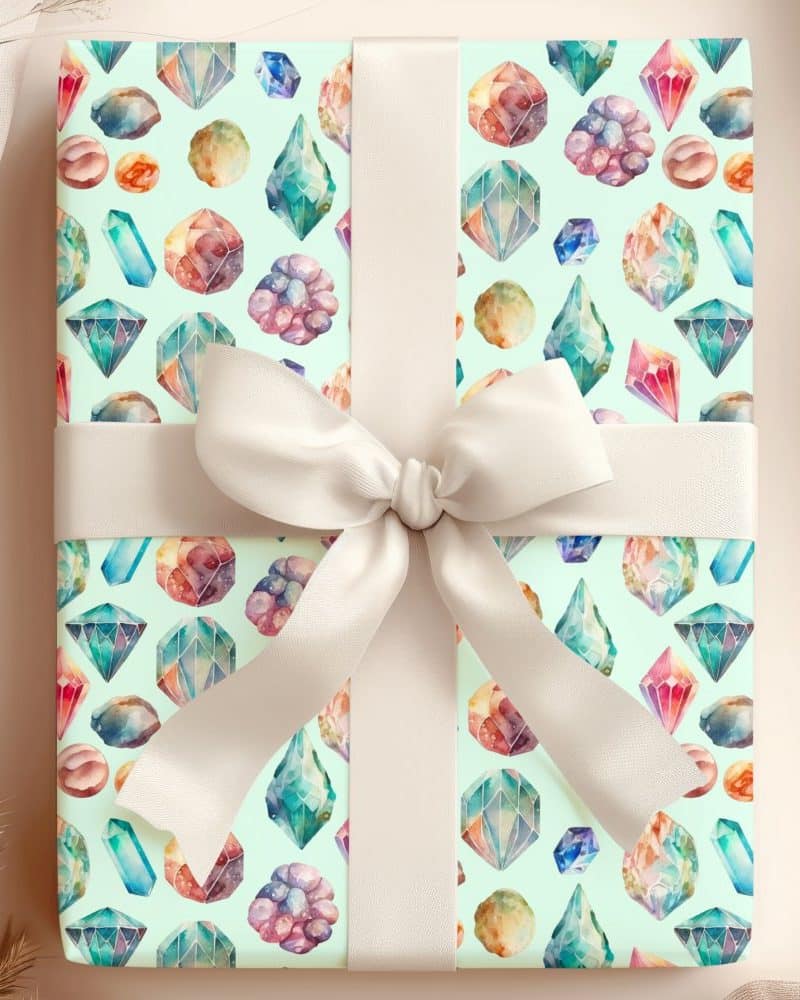 A gift wrapped in light green paper featuring a watercolor pattern of colorful gemstones and crystals, tied with a cream-colored ribbon.