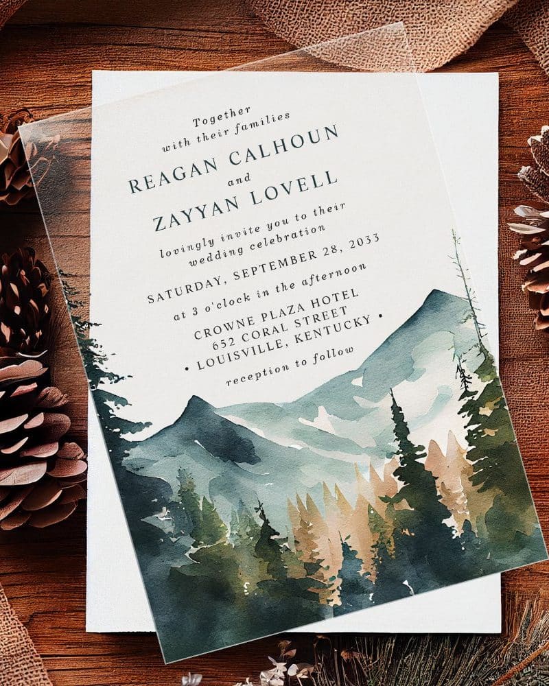 Clear wedding invitation with watercolor mountain and forest scene. The invitation features the names Reagan Calhoun and Zayyan Lovell.