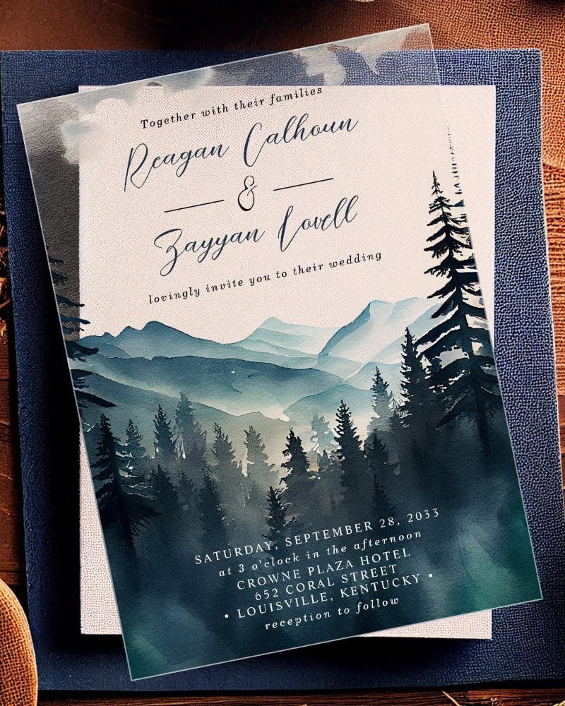 Clear wedding invitation with watercolor painting of mountains and pine trees. The names Reagan Calhoun and Zayyan Lovell are prominently displayed.