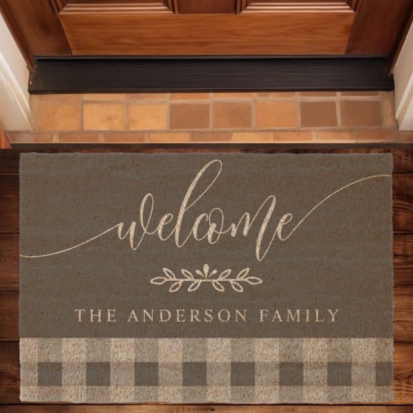 Gray Welcome Mat With The Words Welcome, A Small Floral Design, And The Anderson Family In A Light Beige Script. The Bottom Features A Tan And Gray Gingham Pattern.