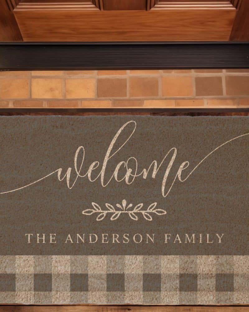Gray welcome mat with the words Welcome, a small floral design, and The Anderson Family in a light beige script. The bottom features a tan and gray gingham pattern.