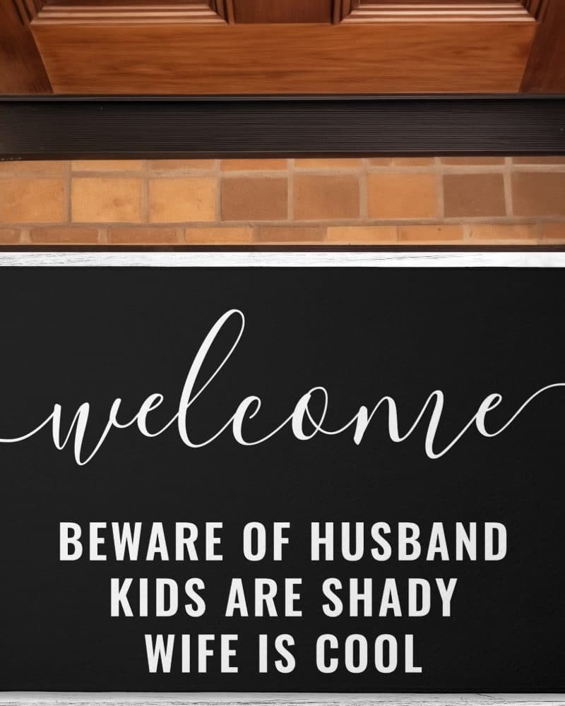 Black welcome mat with white script lettering that says Welcome, Beware of Husband, Kids are Shady, Wife is Cool.