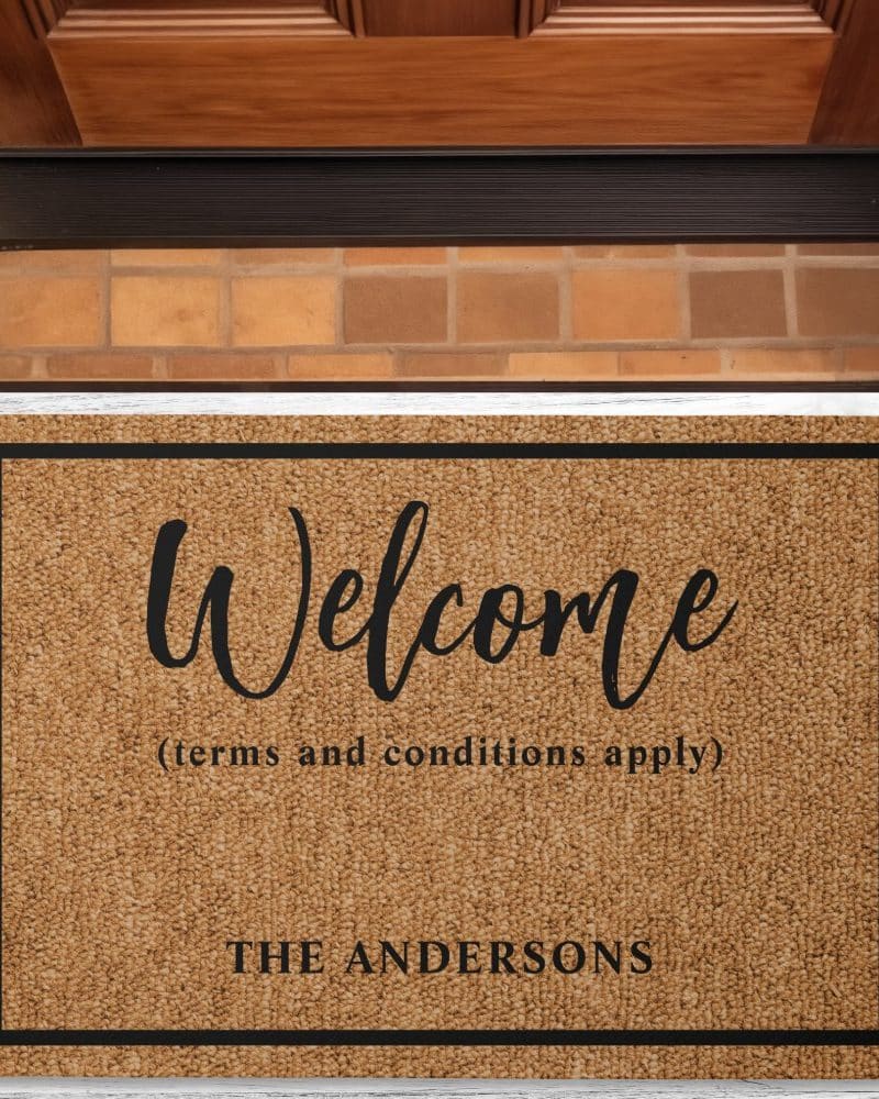 A tan welcome mat with black text. The words Welcome, terms and conditions apply, and The Andersons are visible.