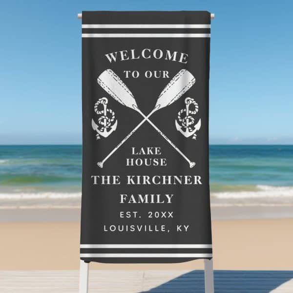 Black Beach Towel With White Lettering That Says Welcome To Our Lake House, The Kirchner Family, Est. 20Xx, Louisville, Ky. Crossed Oars And Anchors Are Depicted Above The Text.