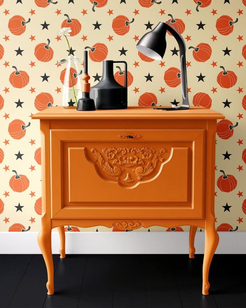 Orange painted dresser with ornate detailing, holding a lamp and decorative objects, against a wall with a pumpkin and star pattern.