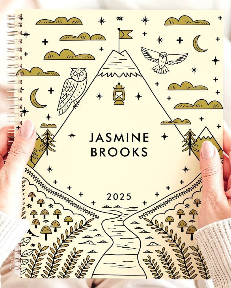 Two hands hold a 2025 planner featuring a cream cover with gold line drawings of mountains, owls, a lantern, and a winding river. The name Jasmine Brooks is printed on the cover.