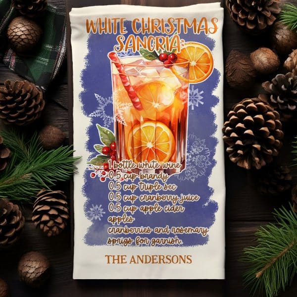 White Christmas Sangria Recipe On A Kitchen Towel, Featuring An Illustration Of A Glass Of Sangria With Oranges And Cranberries.