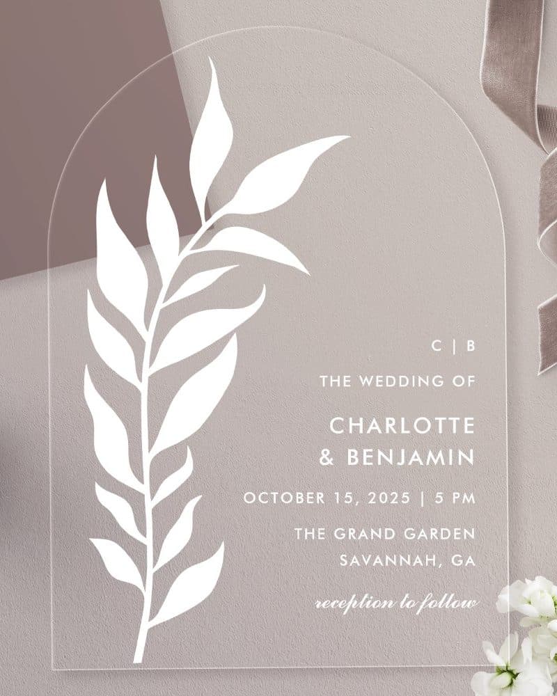 Arch-shaped acrylic wedding invitation with a white stylized leaf design and the couple's names and wedding details.