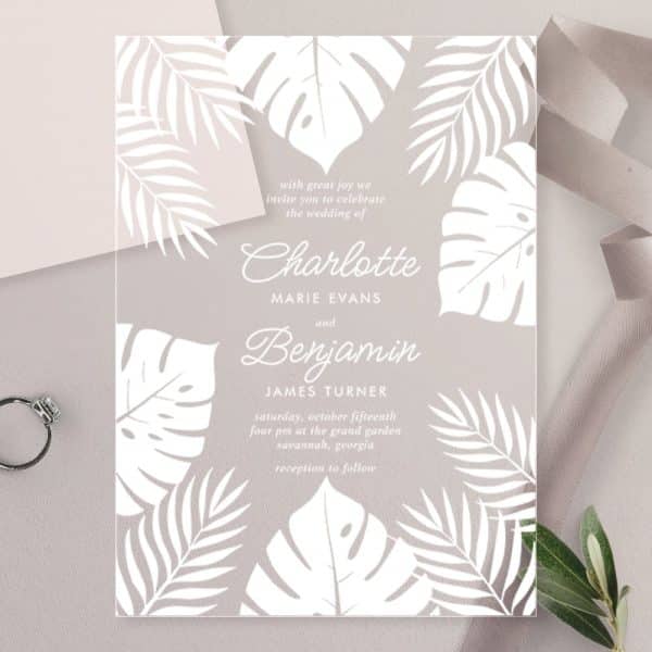 Clear Wedding Invitation With White Tropical Leaves And The Names Charlotte And Benjamin.