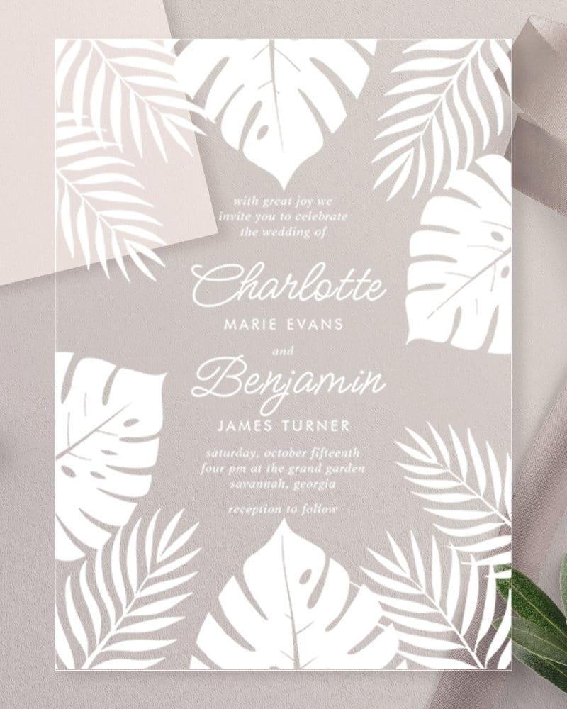 Clear wedding invitation with white tropical leaves and the names Charlotte and Benjamin.