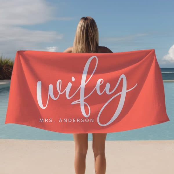 Woman Holding A Coral Beach Towel That Says Wife And Mrs. Anderson In White Script.