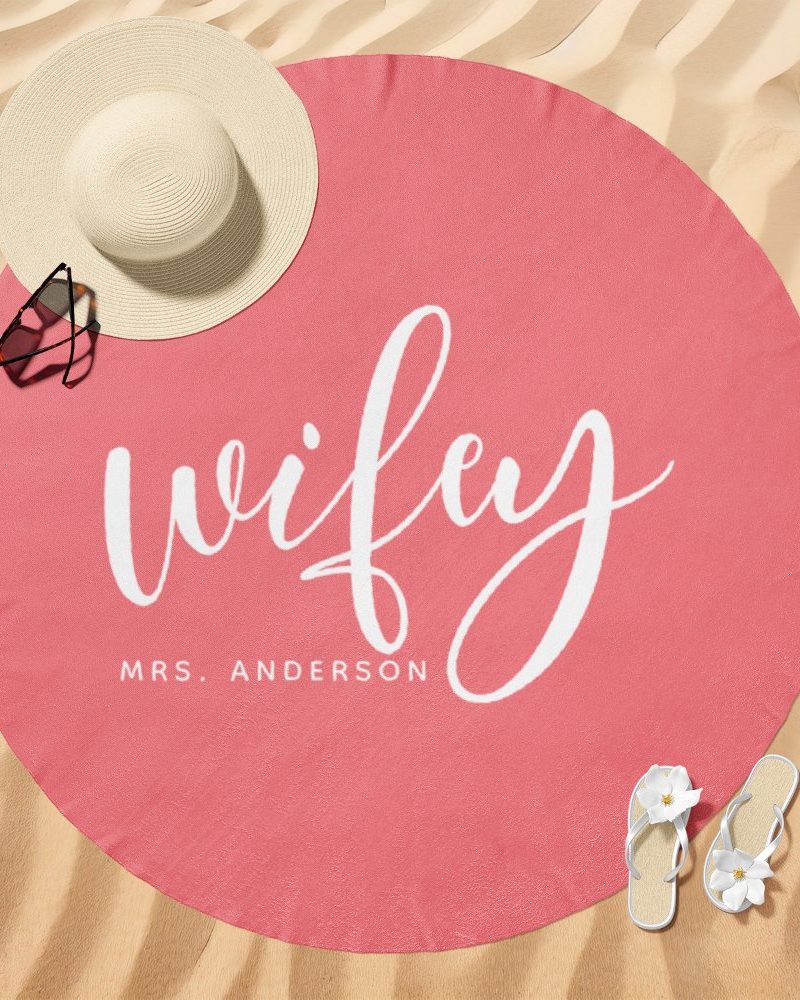Pink round beach towel with the word wifey in white script lettering and Mrs. Anderson below it.