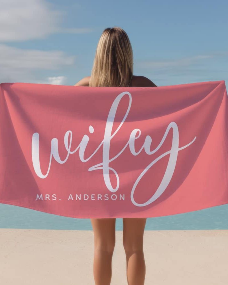 Woman holding coral beach towel with the word wifey in white script lettering and her last name below.