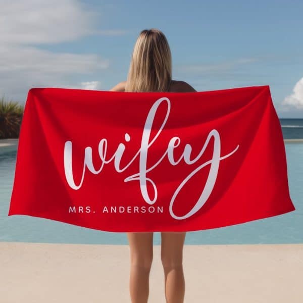 Woman Holding A Red Beach Towel That Says &Quot;Wifey&Quot; And &Quot;Mrs. Anderson&Quot; In White Script Lettering.