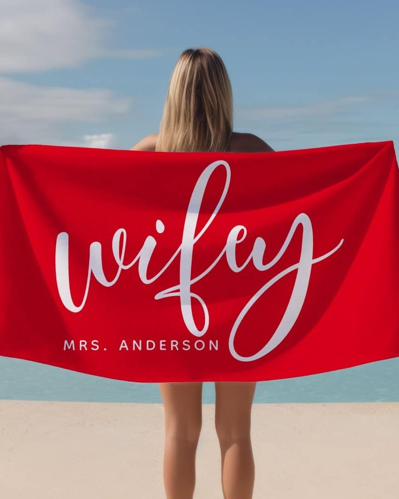Woman holding a red beach towel that says "wifey" and "Mrs. Anderson" in white script lettering.
