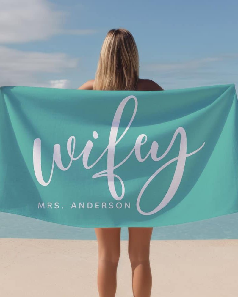 Woman holding a teal beach towel with the word wifey and Mrs. Anderson in white script.