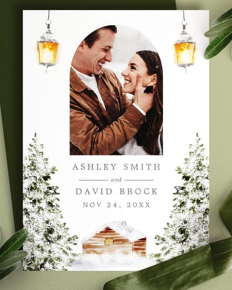 Wedding invitation featuring a photo of a smiling couple, winter-themed artwork including snow-covered trees and a cabin, and the couple's names and wedding date.