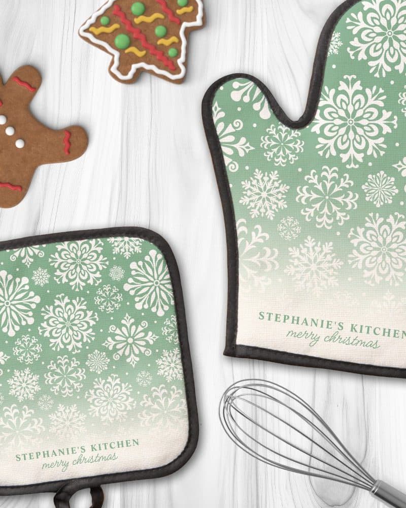 A pair of green oven mitts with a snowflake pattern and personalized Christmas text.
