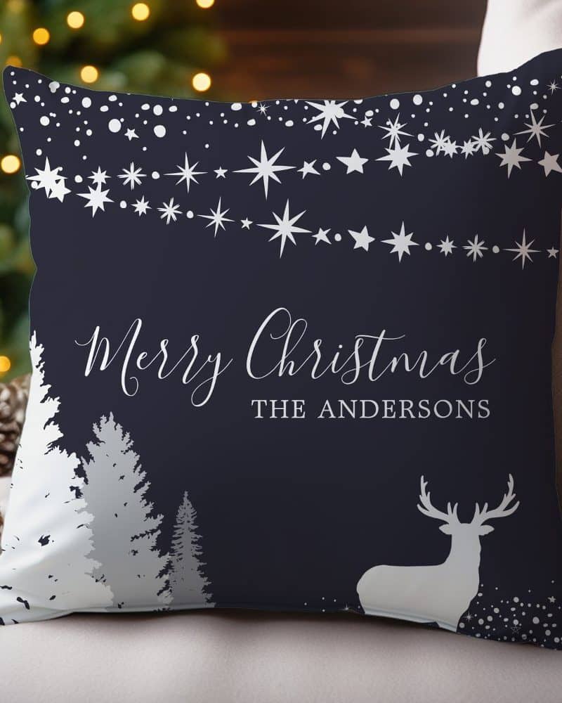 Dark blue throw pillow with a silver Merry Christmas message, the Andersons family name, a deer silhouette, and a snowy forest scene.