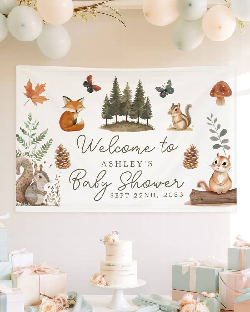 A woodland-themed baby shower banner for Ashley, featuring cartoon animals like foxes and squirrels, along with trees and pinecones. The date is also included.