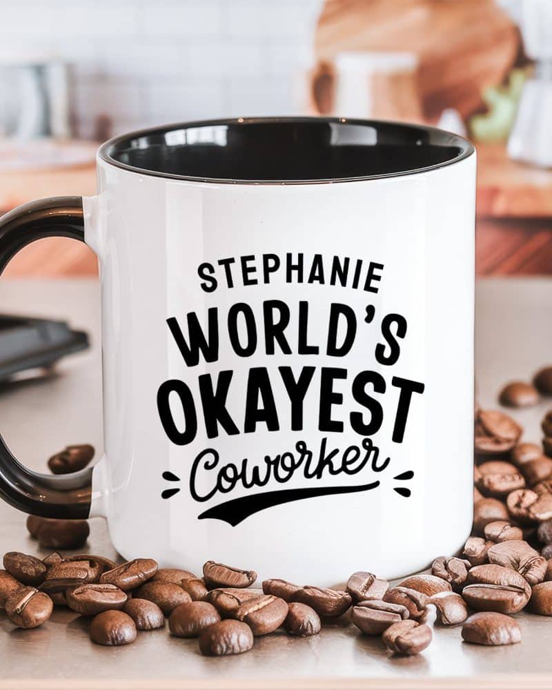 White coffee mug with black handle and interior, displaying the text Stephanie, World's Okayest Coworker.