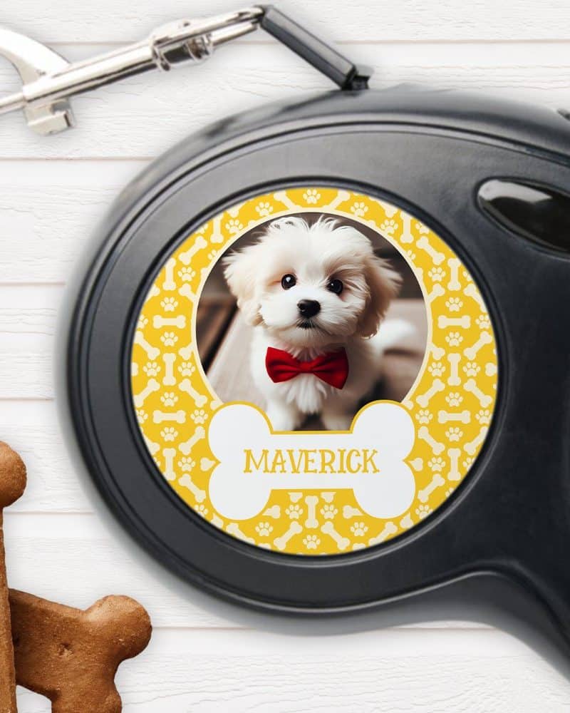 Retractable dog leash with a yellow bone-shaped design featuring a photo of a small white dog wearing a red bow tie and the name Maverick.