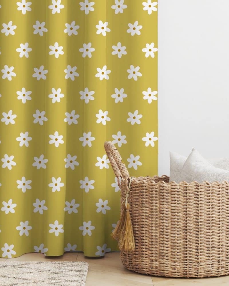 Mustard yellow curtain with a repeating pattern of white daisies, next to a wicker basket containing several white pillows.