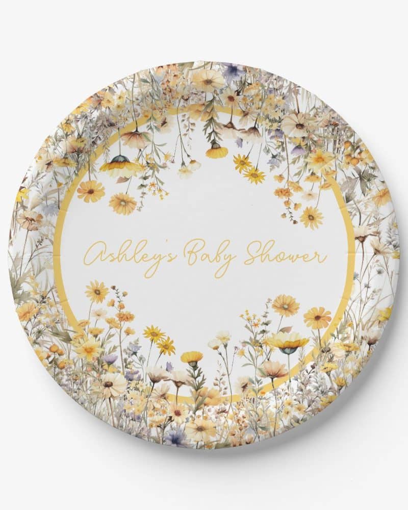 A round paper plate decorated with a ring of yellow and white wildflowers, with Ashley's Baby Shower written in the center.