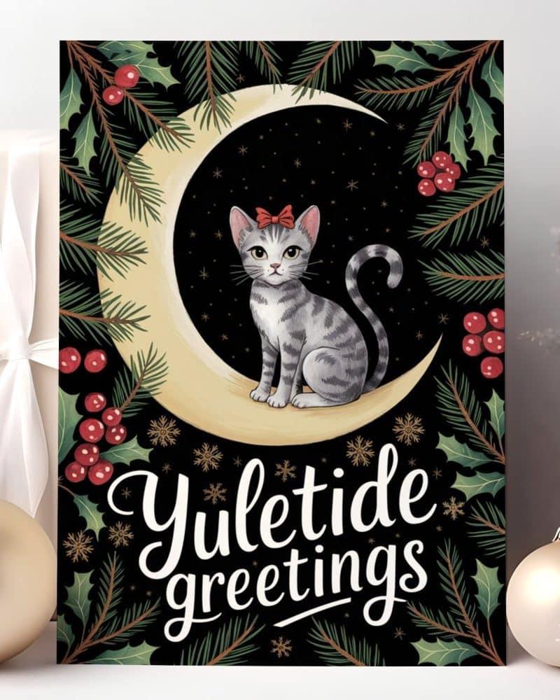 A grey tabby cat wearing a red bow sits on a crescent moon surrounded by holly and pine branches. The words Yuletide Greetings are below.