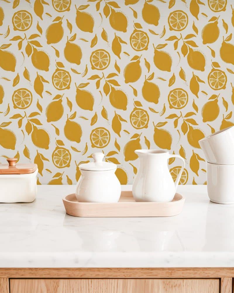 White kitchenware sits on a marble counter in front of a mustard yellow lemon pattern wallpaper.