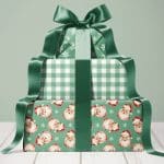 A stack of three Christmas presents wrapped in green paper with varying patterns: snowflakes, gingham, and vintage Santa faces. A large green ribbon ties the stack together.