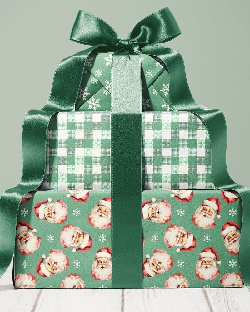 A stack of three Christmas presents wrapped in green paper with varying patterns: snowflakes, gingham, and vintage Santa faces. A large green ribbon ties the stack together.